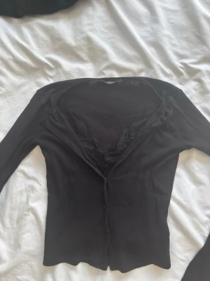 Svart kofta med spets från ZARA - Snygg svart kofta från ZARA med knäppning framtill och spetsdetaljer vid halsringningen. Tunn och figurnära modell med långa ärmar, perfekt att styla över ett linne eller topp. Enkel men elegant design som passar till många outfits.