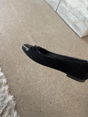 Svarta ballerinaskor med lackdetalj - Stilrena svarta ballerinaskor med glansig lackad tå och söt rosett framtill. Skorna har en klassisk rundad form och dekorativa sömmar längs kanten. Materialet är en mix av mockaimitation och lack, vilket ger en snygg kontrast.
