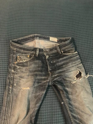 Blå slitna jeans med hål - Säljer ett par blå jeans med slitningar och stora hål på låret och mindre slitningar vid fickorna. Jeansen har en klassisk femficksmodell och tydliga tvätteffekter för en riktigt rå streetlook. Perfekt för dig som gillar en edgy och avslappnad stil.