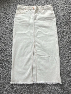 Vit långkjol i denim med slits - Stilren vit långkjol i denim med rå kant nertill och slits framtill. Kjolen har klassiska jeansfickor både fram och bak samt bälteshällor i midjan. Perfekt för dig som gillar en enkel men trendig look. Midjemåttet är ca 34cm. 