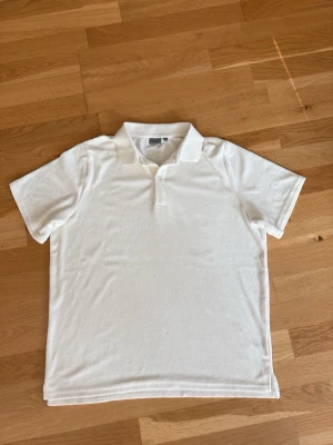 Vit pikétröja med krage - Klassisk vit pikétröja från Ryvls med kort ärm och krage. Tröjan har en enkel knappslå framtill och är gjord i ett mjukt, bekvämt material. Perfekt för en clean och stilren look.