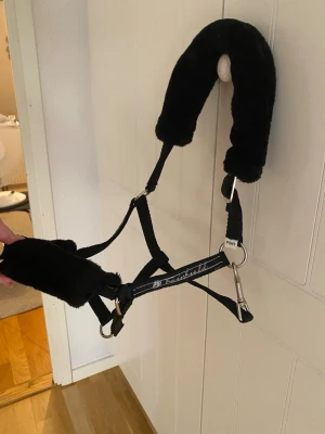 Svart grimma med mjukfodring pony - Svart grimma för ponny med mjuk, fluffig fodring på nack- och nosdel. Tillverkad i slitstarkt nylon med metallspännen och detaljer. Märkt med 'PONY' och har broderad text på sidan. Perfekt för dig som vill ha extra komfort till din häst.