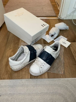 Valentino vita sneakers med blå detalj - Snygga vita sneakers från Valentino Garavani med bred mörkblå rem över vristen. Klassisk låg modell med rund tå och diskret logga på plösen. Tillverkade i läder och levereras med originalkartong och dustbag.storlek 43 passar 42