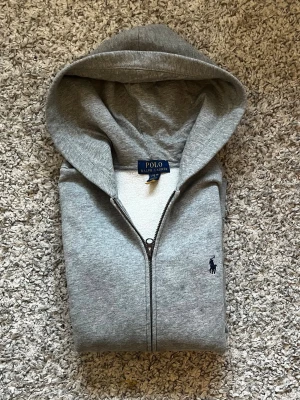 Ralph Lauren Zipup Grå - Ralph Lauren Fleece Full-Zip Hoodie – Grå   Stilren och tidlös Ralph Lauren hoodie i mjuk fleece med klassisk grå färg och det ikoniska Polo Ralph Lauren-loggan på bröstet. Full-zip design med huva och praktiska kängurufickor. Tyget har gått upp en liten bit men det är inte något som märks. Skriv om du vill ha bild.  Perfekt till vardags, skolan eller en clean streetwear-look. Enkel att matcha med jeans, mjukisbyxor eller tracksuit.  ✨ Detaljer: • Polo Ralph Lauren • Mjuk fleece • Full-zip • Huv