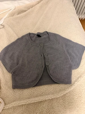 Grå kortärmad stickad kofta H&M ( Stockholm stil inspirerad💓💓) - Kortärmad stickad kofta från H&M i mjuk grå färg. Modellen är croppad med rund halsringning och stora knappar framtill. Perfekt att slänga över en topp för en chill och snygg look. Ribbstickade kanter ger extra detalj.