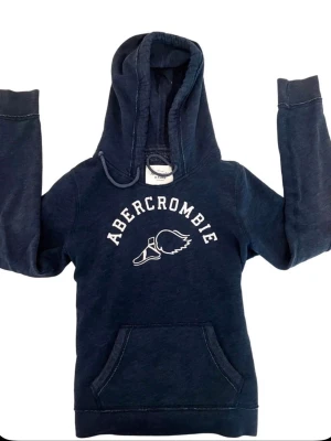 Mörkblå hoodie från Abercrombie - Mörkblå hoodie med vit Abercrombie-logga och älgmotiv på bröstet. Klassisk känguruficka framtill, dragsko i huvan och ribbade muddar. Perfekt för en avslappnad och cool stil.