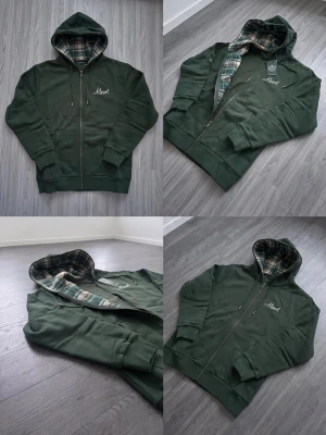 Mörkgrön hoodie med rutigt foder - Snygg mörkgrön hoodie med dragkedja och huva. Insidan av huvan har ett rutigt mönster i grönt, vitt och beige. Broderad text på bröstet. Långärmad modell med ribbade muddar och fickor framtill. Perfekt för dig som gillar en chill och stilren look.