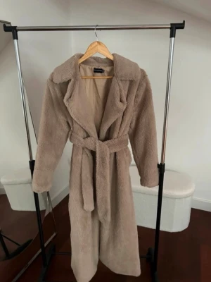 Beige fluffig lång kappa med skärp - Säljer en supermjuk och fluffig beige kappa med bred krage och knytskärp i midjan. Kappan är lång och har en oversized look som ger en riktigt trendig vibe. Perfekt för dig som vill ha en cozy men ändå stylish ytterjacka till hösten eller vintern.