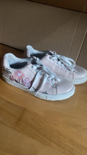 Rosa sneakers med broderade blommor - Snygga sneakers med färgglada broderade blommor på sidan. Skorna har vita snören och en klassisk låg design. Perfekta för dig som vill ha en unik och trendig look med lite extra detaljer.