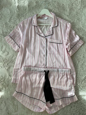 Pyjamas - Söt pyjamas i ljusrosa och vita ränder med svarta kontrastsömmar. Setet består av en kortärmad skjorta med bröstficka och matchande shorts med svart knytband i midjan. Tillverkad i ett mjukt, glansigt material som känns lyxigt mot huden.