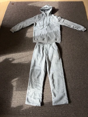 Grått mjukisset med hoodie och byxor - Säljer ett grått mjukisset med dragkedja och huva på tröjan samt två fickor framtill. Byxorna har resår i midjan och raka ben. Båda delarna har ett broderat blommönster i vitt. Perfekt för chill dagar hemma eller till träningen.