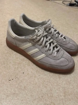 Adidas Handball Spezial grå/vita sneakers - Snygga Adidas Samba OG sneakers i grå mocka med vita detaljer och klassiska tre ränder på sidan. Skorna har vit hälkappa, vita skosnören och en brun gummisula. Perfekt för dig som gillar retrostil och streetwear.