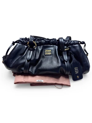 Svart handväska i läder från Miu Miu - Snygg svart handväska från Miu Miu Archive 2006 i mjukt läder. Väskan har guldfärgade detaljer, dragsko upptill och en tag med logga. Perfekt för dig som gillar exklusiva accessoarer med vintagekänsla. Nypris 35 000kr