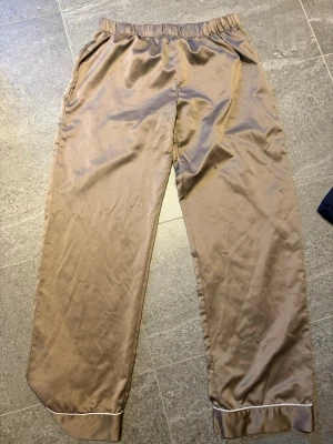 Bruna satinpyschbyxor i storlek M - Snygga och bekväma bruna loungewear-byxor från Lindex i satinmaterial. Byxorna har resår i midjan, raka ben och vita passpoaldetaljer vid bensluten. Perfekta för chill dagar hemma eller mysiga kvällar.