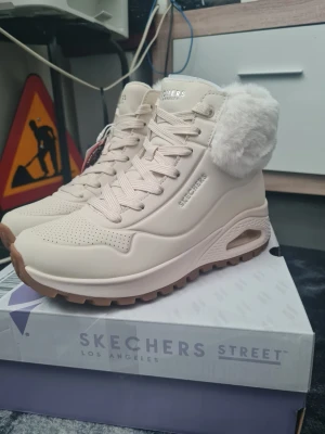Beige kängor med fuskpäls från Skechers stl 38 - Säljer ett par snygga beige kängor från Skechers med chunky sula och vit fuskpäls runt skaftet. Kängorna har perforerade detaljer, grova snören och en stabil yttersula i gummi. Perfekta för kalla dagar när du vill ha både stil och komfort. Helt nya och oanvända i kartong! Stl 38