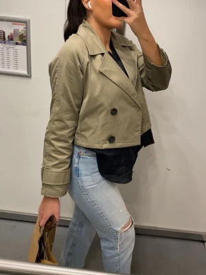 Kort trenchcoat från ZARA  - Snygg kort trenchcoat från ZARA i khakigrönt bomullstyg. Passar XS-M, jackan har dubbelknäppning med stora svarta knappar, bred krage och uppvikta ärmslut. Perfekt för en trendig och avslappnad look till vardags.