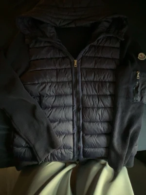 Mörkblå pufferjacka från Moncler - Snygg mörkblå pufferjacka från Moncler med quiltad front, stickade ärmar och huva. Jackan har dragkedja framtill och en Moncler-logga på ärmen. Perfekt för kalla vinterdagar och ger en sportig men ändå stilren look.