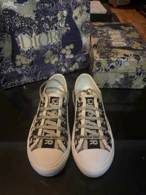 Dior sneakers med monogrammönster - Snygga låga sneakers från Dior med svartvitt monogrammönster och vita sulor. Skorna har vita snören och tydlig 'Christian Dior'-logga på plösen. Tillverkade i canvas med klassisk siluett och trendig design som sticker ut.