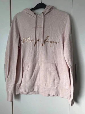 Ljusrosa hoodie med guldtext - Ljusrosa hoodie med huva och dragsnören. Framtill står det 'Magnifique' i glansig guldtext. Hoodien har en stor magficka och ribbade muddar vid ärmar och nederkant. Perfekt för en chill och avslappnad stil.