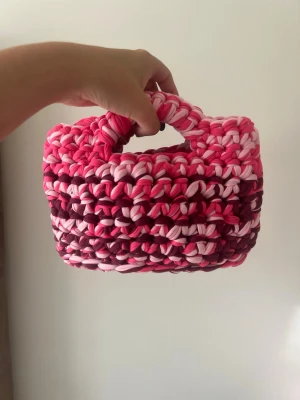 Handvirkad rosa handväska - Unik handväska som är handvirkad i tjockt garn med olika nyanser av rosa och vinrött. Väskan har en rektangulär form och ett fast handtag, perfekt för dig som vill sticka ut med en färgglad accessoar.💕