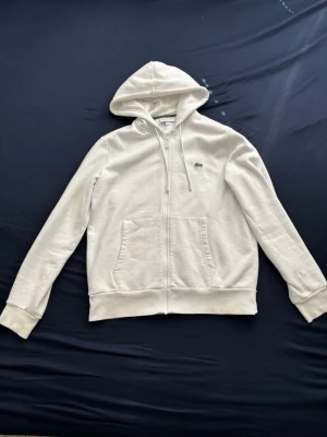 Vit zip-hoodie från Lacoste - Snygg vit hoodie med dragkedja från Lacoste. Klassisk krokodil-logga på bröstet, två stora fickor fram och ribbade muddar. Tillverkad i mjukt material och har en justerbar huva med snören. Perfekt för en clean och avslappnad stil.