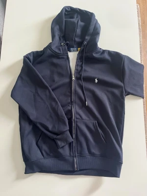 Marinblå hoodie Ralph Lauren - Snygg marinblå hoodie från Polo Ralph Lauren med dragkedja. Tillverkad i mjukt bomullsmaterial och har fickor framtill. Perfekt för en avslappnad och stilren look.