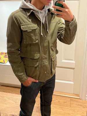 Olivgrön Fieldjacket från Native Youth - Säljer en lite tunnare, snygg olivgrön Fieldjacket från Native Youth, med flera praktiska fickor framtill och tryckknappar. Passar perfekt att använda lager-på-lager nu inför hösten. Storlek M. Skriv gärna vid minsta fundering! 