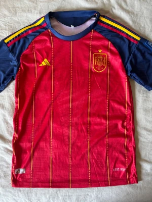 Spaniens matchtröja röd/blå Adidas - Säljer en officiell matchtröja för Spaniens landslag från Adidas. Tröjan är röd med gula vertikala ränder, blå ärmar och gula/röda detaljer på axlarna. Broderad landslagssköld och Adidas-logga på bröstet. Tillverkad i lätt funktionsmaterial.