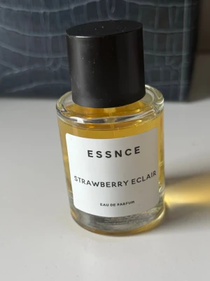 ESSNCE Strawberry Eclair parfym - ESSNCE Strawberry Eclair Eau de Parfum i en rund 50 ml glasflaska med svart lock. Parfymen har en ljusgul vätska och en stilren vit etikett med svart text. Doften har troligen söta och fruktiga toner av jordgubbe och bakverk.