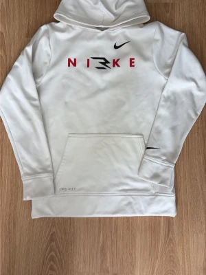 Vit hoodie med tryck från Nike - Vit hoodie från Nike med röd och svart logga och text på bröstet. Klassisk känguruficka framtill och Dri-FIT-material som andas. Långärmad med huva och diskret Nike-logga på ärmen. Perfekt för en sportig och avslappnad stil.