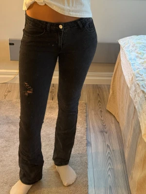 Svarta bootcut jeans med slitning - Säljer ett par svarta bootcut jeans med slitning på vänster lår. Jeansen har klassisk femficksmodell, normal midja och stängs med knapp och dragkedja. Materialet är denim med lite stretch för skön passform.
