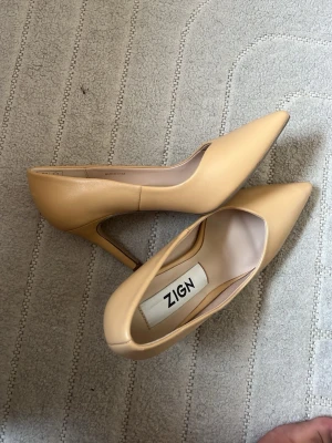 Beige pumps med smal klack från Zign - Stilrena pumps i beige från Zign med spetsig tå och smal klack. Skorna är tillverkade i ett slätt material som ger en elegant look. Perfekta för dig som vill ha en klassisk och trendig sko till garderoben.