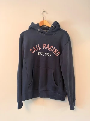 Mörkblå Sail Racing hoodie - Mörkblå hoodie från Sail Racing med trycket 'SAIL RACING EST. 1977' i vitt och rött på bröstet. Klassisk känguruficka framtill och huva med dragsko. Mjuk bomullsmix och ribbade muddar vid ärmslut och nederkant. Perfekt för en avslappnad stil.