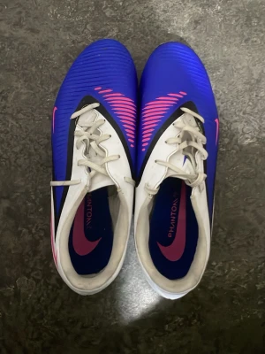 Nike Phantom GT2 blå fotbollsskor - Säljer ett par Nike Phantom GT2 fotbollsskor i blått med vita och rosa detaljer. Skorna har snörning, syntetiskt material och en modern design med rosa Nike-logga och mönster på ovandelen. Perfekta för dig som vill sticka ut på planen.