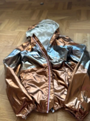 Metallic vindjacka från JOTT - Snygg vindjacka från JOTT i metallic koppar och silver med huva och dragkedja. Jackan har elastiska muddar vid ärmslut och nederkant samt ett coolt tryck på insidan. Perfekt för dig som vill sticka ut med en unik look.