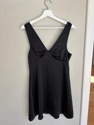 Svart satin klänning med v-ringning - Snygg svart klänning i satin med breda axelband och djup v-ringning både fram och bak. Klänningen har en lätt utställd passform och slät yta som ger en elegant look. Perfekt för dig som gillar minimalistisk och stilren design.