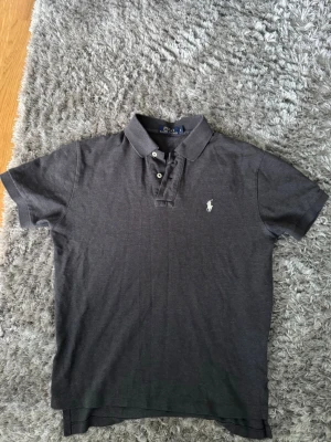 Mörkgrå pikétröja från Polo Ralph Lauren - Klassisk mörkgrå pikétröja från Polo Ralph Lauren med korta ärmar och vit broderad logga på bröstet. Tröjan har krage och knäppning med två knappar framtill. Perfekt till en avslappnad men ändå stilren look.