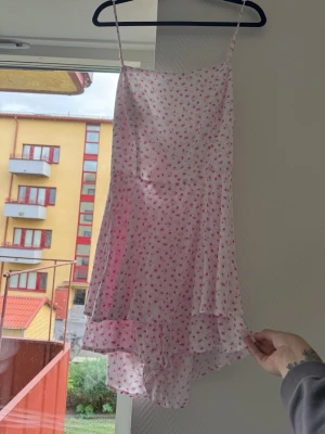 Vit blommig klänning med inbyggda shorts - Söt vit kortklänning med rosa blommönster och tunna axelband. Klänningen har volangdetalj nedtill och är tillverkad i ett lätt, glansigt material som ger en luftig känsla. Perfekt för varma dagar och somriga tillfällen.