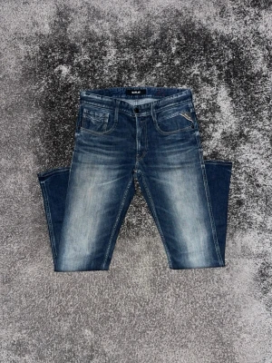 Blå slitna jeans med tidlös tvätt från Replay  - Säljer ett par blå jeans med snyggt slitna detaljer från Replay. Jeansen har klassisk femficksmodell, knappgylf och kontrastsömmar. Perfekta för en avslappnad och trendig look.