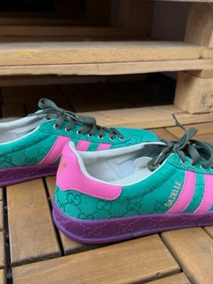 Gucci x Adidas Gazelle gröna sneakers - Unika Gucci x Adidas Gazelle sneakers i grönt tyg med rosa detaljer och lila sula. Skorna har mörkgröna skosnören, klassiska Adidas-ränder och GG-mönster över hela ovandelen. Insidan är vit och märkt med både Gucci och Adidas. Perfekt för dig som vill sticka ut.
