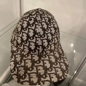 CD keps - Säljer en CD keps med svart och beige all-over logotypmönster. Kepsen har böjd skärm och klassisk sexpanelsdesign. Materialet ser ut att vara vävt tyg med tydlig struktur. Perfekt för dig som vill sticka ut med en trendig accessoar.