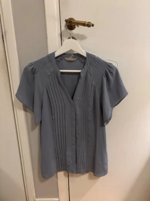 Ljusblå blus med veckad front - Snygg ljusblå blus med korta ärmar och dekorativa veck framtill. Blusen har en avslappnad passform och rundad halsringning. Perfekt att matcha med både jeans och kjol för en trendig look. Lite stor i storleken.