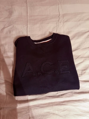 Svart sweatshirt från WHO's A.C.E - Svart sweatshirt med broderad text 'A.C.E' framtill. Klassisk rund halsringning och ribbade muddar. Tillverkad i mjukt material och har en diskret, stilren look med en liten detalj i rött och vitt på insidan av kragen.