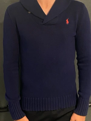 Mörkblå stickad tröja Ralph Lauren - Snygg mörkblå stickad tröja från Ralph Lauren Sport med röd logga på bröstet. Tröjan har sjalkrage med knappdetalj, långa ärmar och ribbade muddar. Tillverkad i 100% bomull för en skön och mjuk känsla.