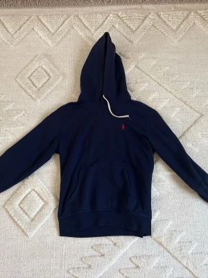 Mörkblå Ralph Lauren hoodie - Snygg mörkblå hoodie från Polo Ralph Lauren med vita snören och klassisk röd logga broderad på bröstet. Hoodien har känguruficka framtill och ribbade muddar vid ärmslut och nederkant. 100% äkta kan fixa digitalt kvitto om beviljat. Haft i ca 2 år med bara använd max 20 gånger med 1 tvätt. Hör av er vid intresse kan skicka bild med den på.