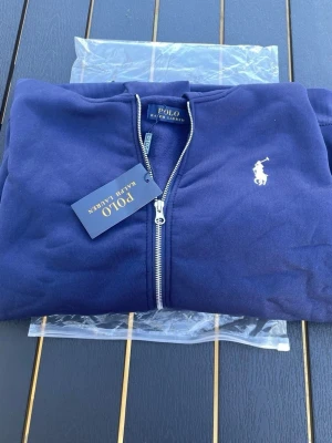 Marinblå hoodie från Polo Ralph Lauren - Snygg marinblå hoodie från Polo Ralph Lauren med dragkedja framtill och den klassiska vita logon broderad på bröstet. Tillverkad i mjukt material och har ribbade muddar vid ärmslut och nederkant. Perfekt för en chill och stilren look.