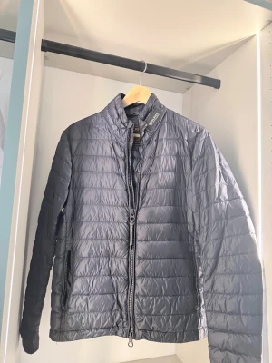  dunjacka från Woolrich  - Snygg  dunjacka från Woolrich i storlek s Jackan har quiltad design, hög krage och dragkedja framtill. Tillverkad i lätt och glansigt material som håller värmen utan att kännas tung. Perfekt för kalla vinterdagar.
