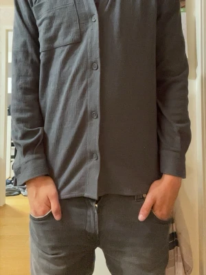 Grå långärmad overshirt/skjorta med ficka - Säljer en stilren grå skjorta med lång ärm och en bröstficka. Skjortan har klassisk krage och knäppning framtill med fyra knappar. Perfekt för dig som gillar en enkel och clean look.