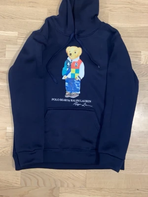 Mörkblå hoodie Polo Bear Ralph Lauren - Mörkblå hoodie från Ralph Lauren med Polo Bear-tryck på bröstet. Klassisk huva med dragsko och stor magficka. Trycket visar en björn i färgglada kläder. Mjuk bomullskänsla och ribbade muddar vid ärmar och nederkant.