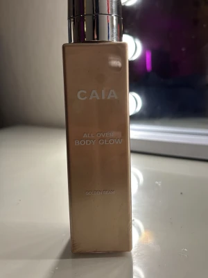 CAIA All Over Body Glow Golden Beam - CAIA All Over Body Glow i nyansen Golden Beam. Kommer i en rektangulär, guldig flaska med pump. Ger huden en snygg glow och skimrande finish. Perfekt för att highlighta kroppen och skapa en solkysst look.